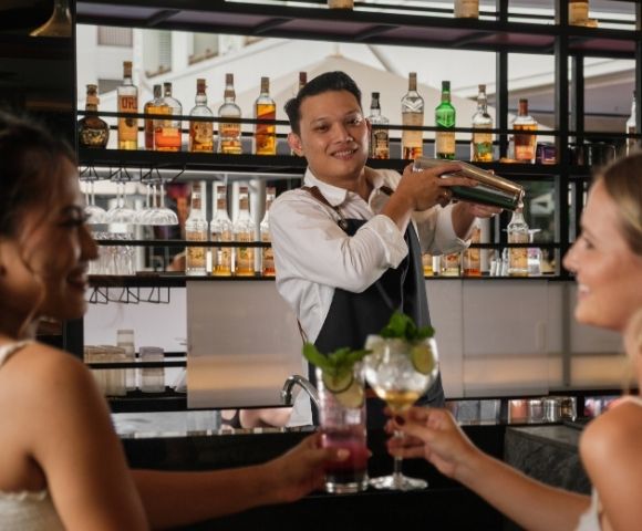 A bartender shakes a cocktail behind a bar while two women enjoy drinks and smile.