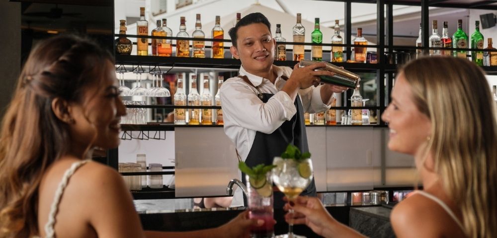 A bartender shakes a cocktail behind a bar while two women enjoy drinks and smile.