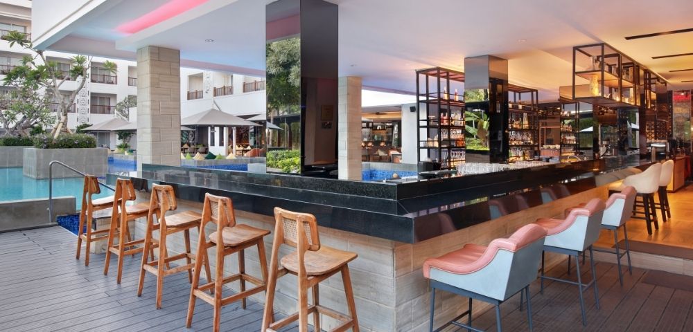 A modern bar area with high stools, located near a pool and surrounded by greenery and seating.