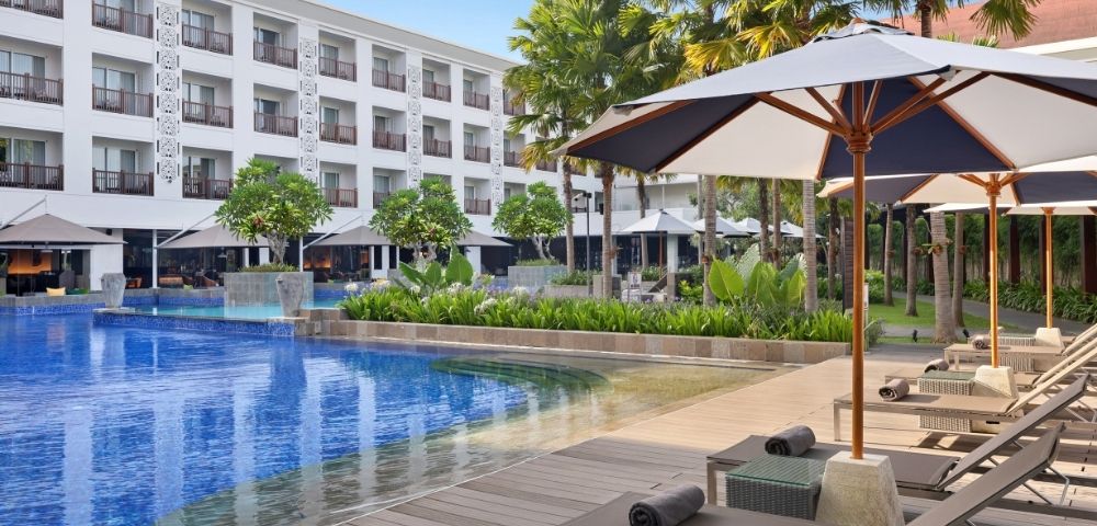 A luxurious hotel pool area with lounge chairs and umbrellas, surrounded by greenery and trees.