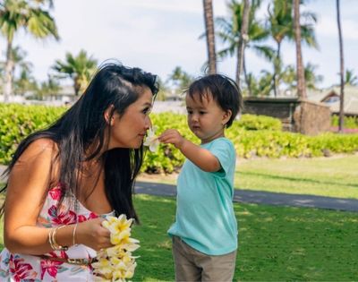 A woman and a young child smell a plumeria flower on a sunny day in a tropical garden with palm trees and lush greenery.