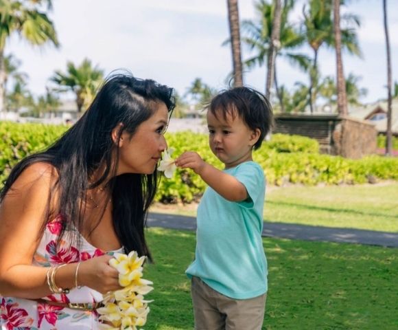 A woman and a young child smell a plumeria flower on a sunny day in a tropical garden with palm trees and lush greenery.