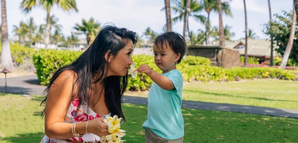 A woman and a young child smell a plumeria flower on a sunny day in a tropical garden with palm trees and lush greenery.