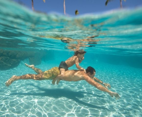 Underwater shot of an adult swimming with a child on their back in clear turquoise water with sunlight dappling on the sandy bottom.