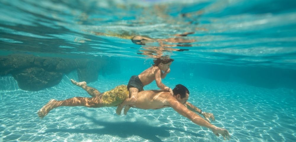 Underwater shot of an adult swimming with a child on their back in clear turquoise water with sunlight dappling on the sandy bottom.