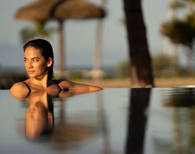 Woman with wet hair wearing a black swimsuit, looking to the side with a relaxed expression, partially submerged in an infinity pool at sunset.