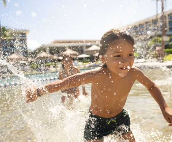A young child runs joyfully through shallow water, splashing with excitement in front of a resort pool.