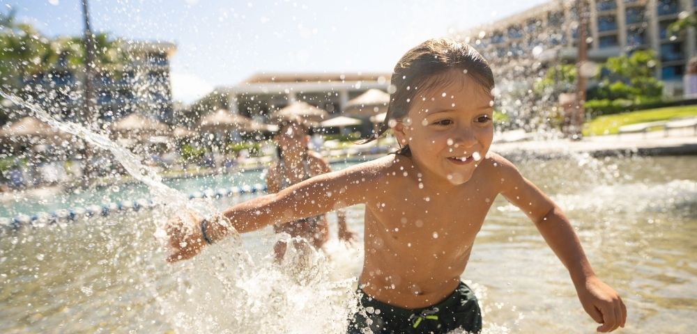 A young child runs joyfully through shallow water, splashing with excitement in front of a resort pool.