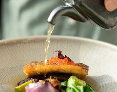 A stream of clear broth is poured from a metal teapot onto a gourmet dish, featuring a seared piece of fish topped with bright orange roe and delicate purple garnishes, presented in a textured ceramic bowl.