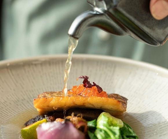 A stream of clear broth is poured from a metal teapot onto a gourmet dish, featuring a seared piece of fish topped with bright orange roe and delicate purple garnishes, presented in a textured ceramic bowl.