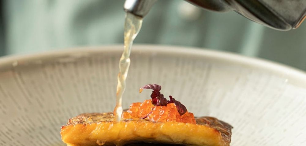 A stream of clear broth is poured from a metal teapot onto a gourmet dish, featuring a seared piece of fish topped with bright orange roe and delicate purple garnishes, presented in a textured ceramic bowl.