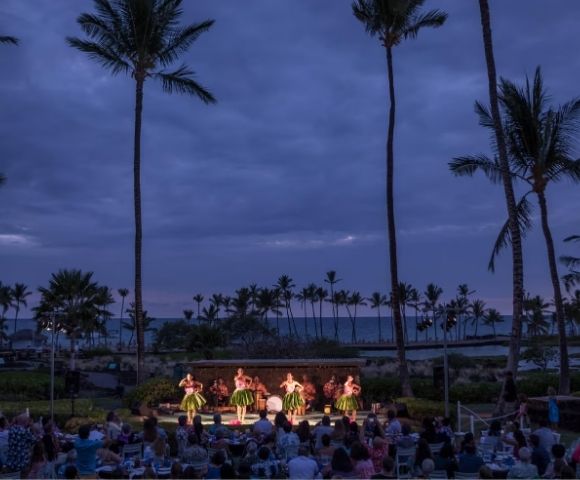 A group of women in grass skirts dance on a stage at dusk, with palm trees silhouetted against a cloudy sky and an audience seated in front of them.