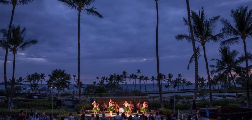A group of women in grass skirts dance on a stage at dusk, with palm trees silhouetted against a cloudy sky and an audience seated in front of them.