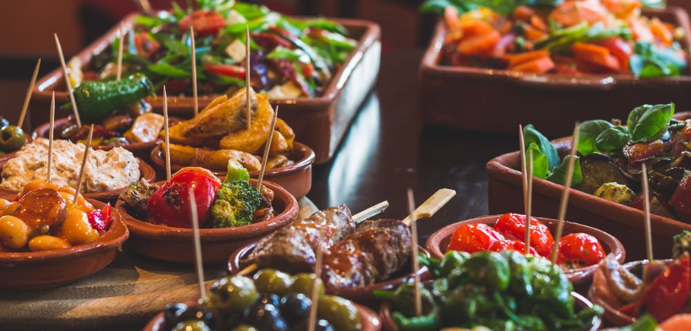 Colorful tapas and pintxos fill rustic dishes, with skewered olives, peppers, vegetables, bread, and savory bites arranged for sharing.