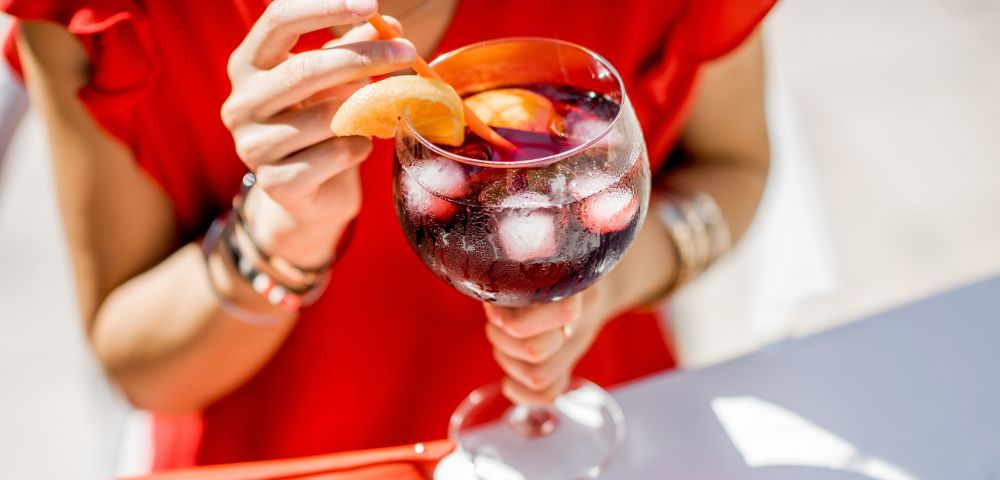 Person holds a sangria with ice and orange garnish, beside a red folding fan in bright sunlight.