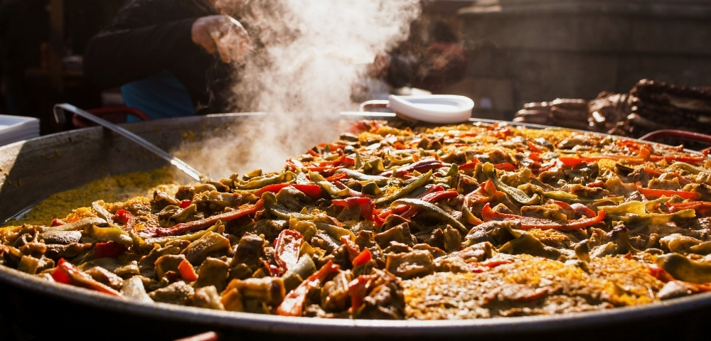 Steaming Spanish paella brimming with rice, peppers, and vegetables fills a market pan as a cook serves it, creating a warm, lively mood.