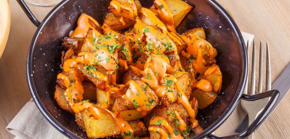 Close-up of crispy roasted potatoes in a black bowl, drizzled with creamy orange sauce and sprinkled with parsley, warm and appetizing.