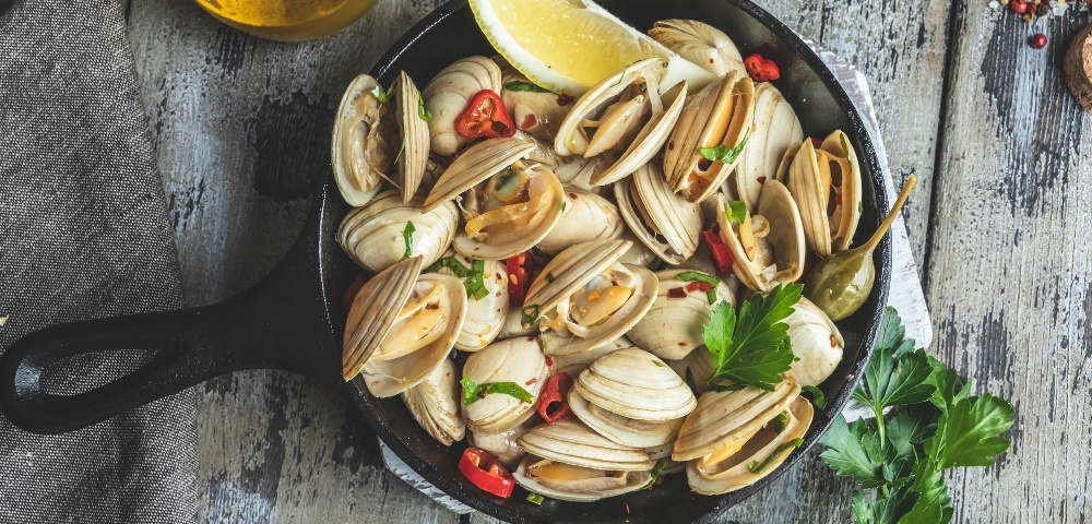 Overhead view of a black skillet filled with steamed clams, lemon wedges, red chili, herbs, and garlic on a rustic wooden table.