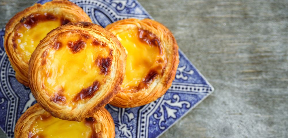 Golden Portuguese custard tarts with flaky, browned pastry sit on a blue patterned plate against a rustic gray table, creating a warm, appetizing mood.