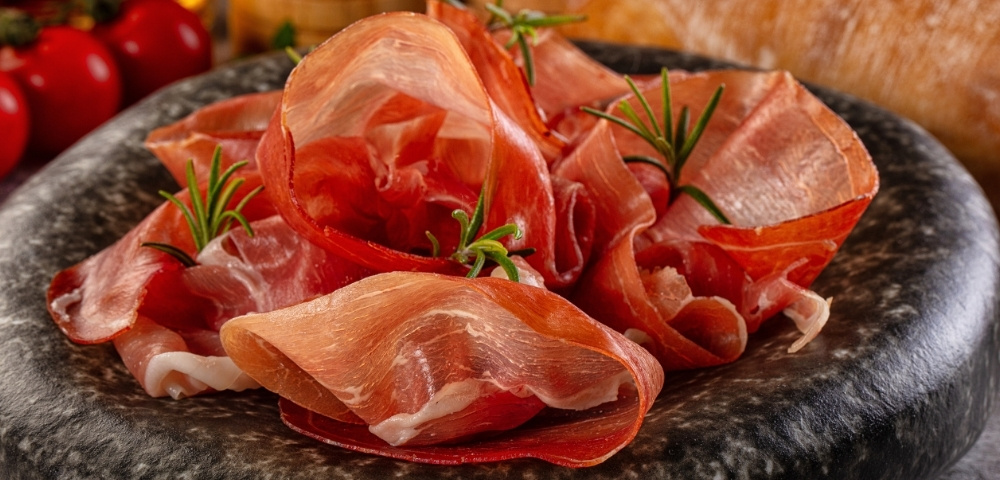 Thinly sliced prosciutto arranged on a dark slate platter with rosemary, tomatoes, and bread, creating a rustic, appetizing spread.