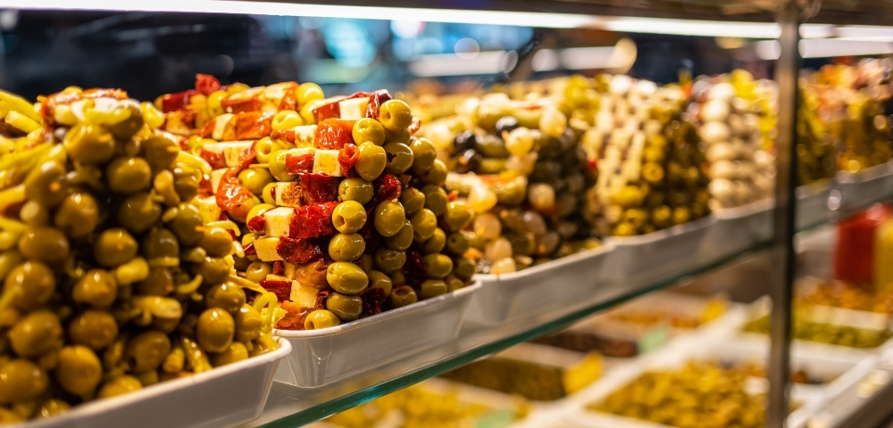 Colorful deli counter displays bowls of green olives, red peppers, and marinated antipasti, creating a warm, inviting market atmosphere.