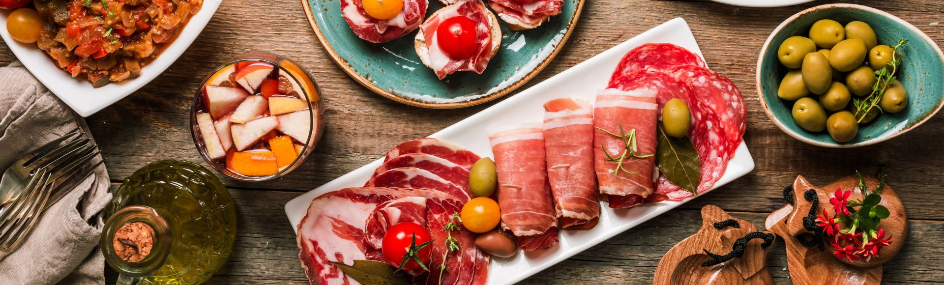 Rustic charcuterie spread with cured meats, olives and tomatoes on colorful plates over a wooden table.