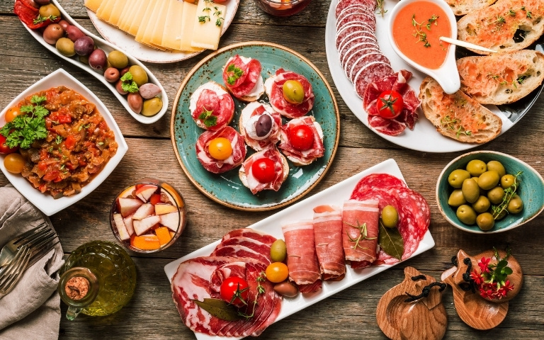 Rustic charcuterie spread with cured meats, olives and tomatoes on colorful plates over a wooden table.