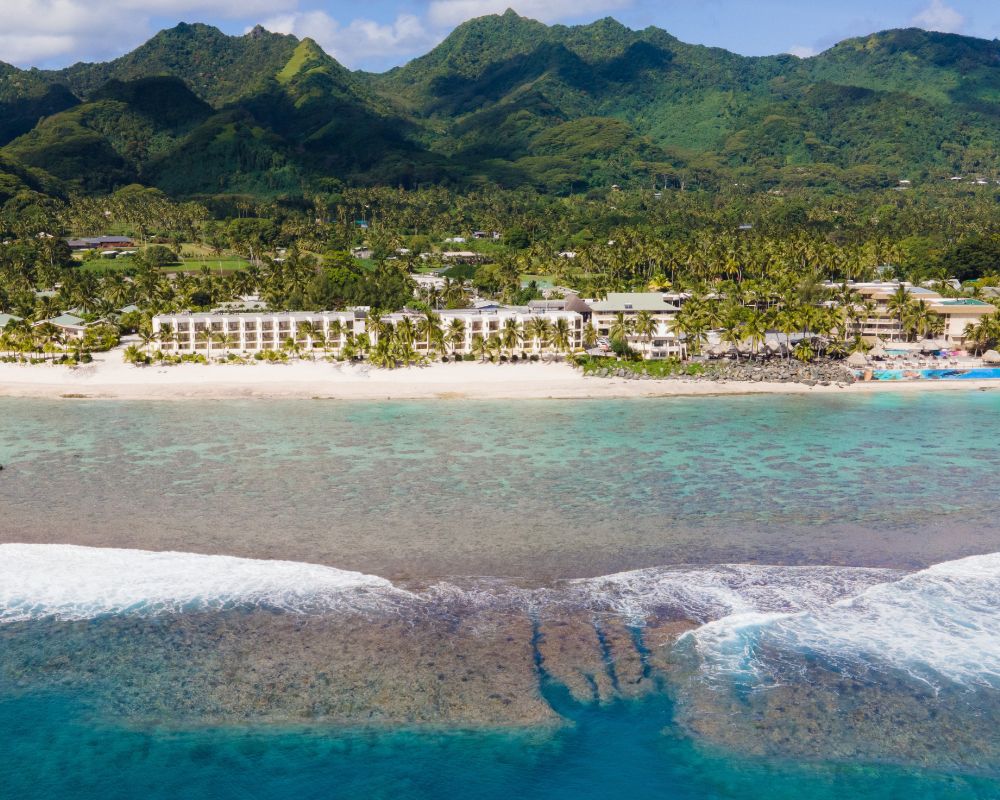 Aerial view of a tropical resort hotel on a white sand beach, lined with palm trees, with turquoise water, coral reefs, and lush green mountains in the background.