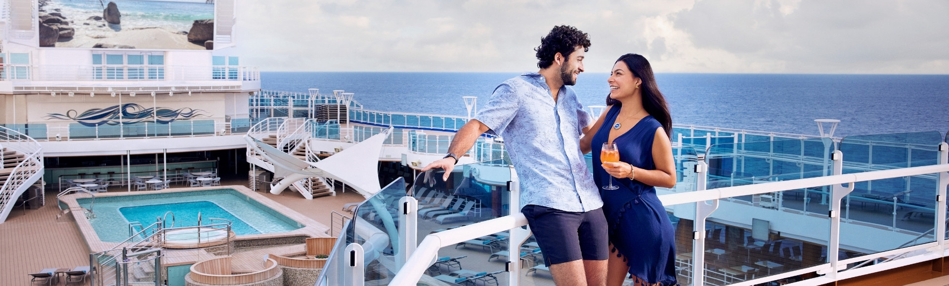 Smiling couple on a cruise ship deck overlooking the blue ocean, holding drinks and enjoying a bright, relaxed vacation moment.