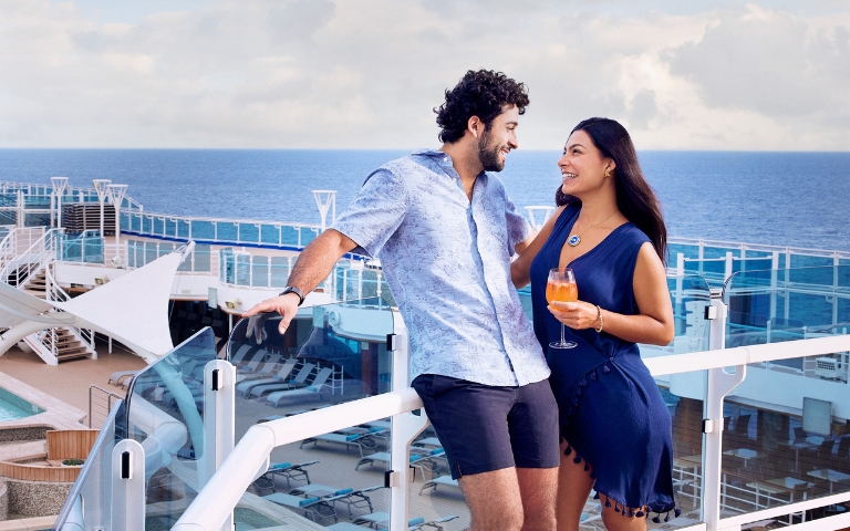 Smiling couple on a cruise ship deck overlooking the blue ocean, holding drinks and enjoying a bright, relaxed vacation moment.