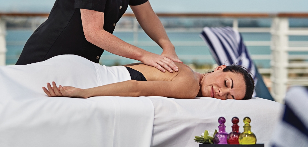 Relaxed woman receives a back massage on a white spa table outdoors; colorful oil bottles sit nearby, creating a calm, soothing atmosphere.