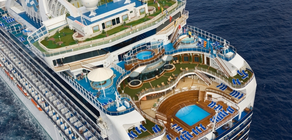 Aerial view of a luxury cruise ship’s blue-and-white decks, pools, water slide, loungers, and sports court surrounded by deep ocean.
