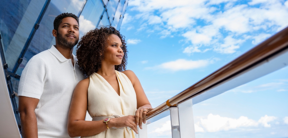 Smiling couple stands on a modern glass balcony, gazing toward a bright blue sky and clouds, conveying warmth, connection, and relaxation.