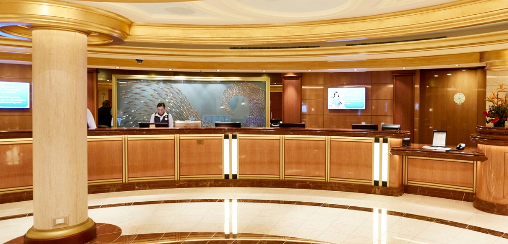 Elegant hotel reception desk with a staff member, rich wood paneling, gold accents, marble floors, and a calm, luxurious atmosphere.