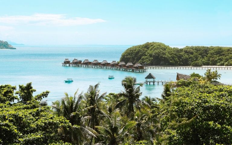 Overwater bungalows with thatched roofs are situated on stilts over turquoise water, with lush green islands and palm trees in the foreground.
