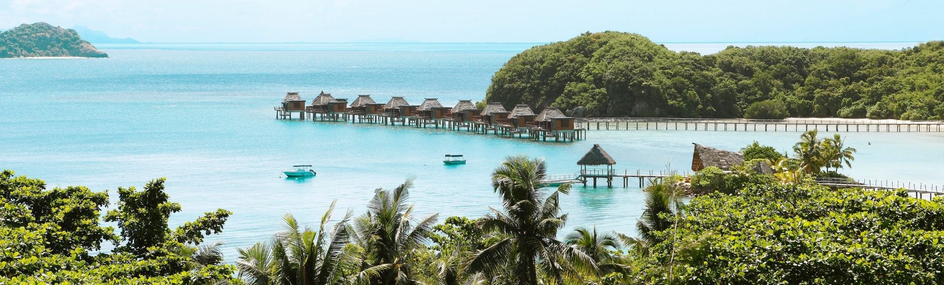 Overwater bungalows with thatched roofs are situated on stilts over turquoise water, with lush green islands and palm trees in the foreground.