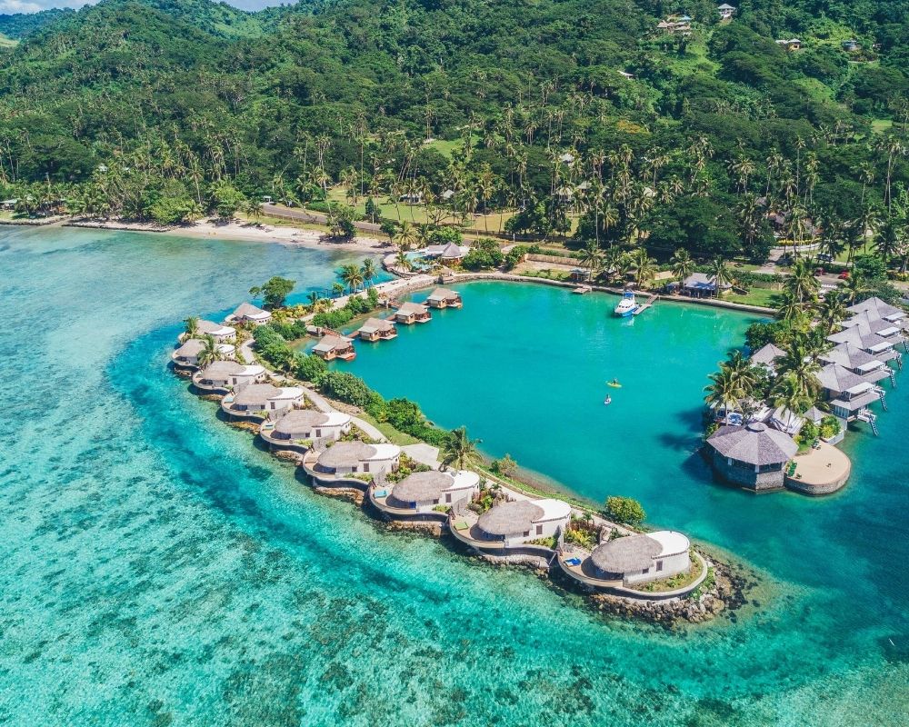 Aerial view of a luxurious resort with overwater bungalows and beachfront villas nestled along a turquoise lagoon with lush green palm trees.