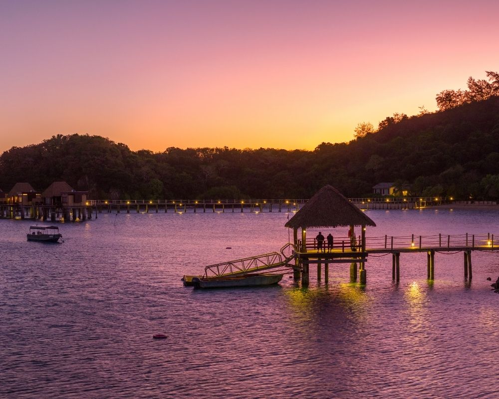Panoramic view of an overwater bungalow resort at sunset, with overwater bungalows and a thatched-roof structure illuminated on a small island.