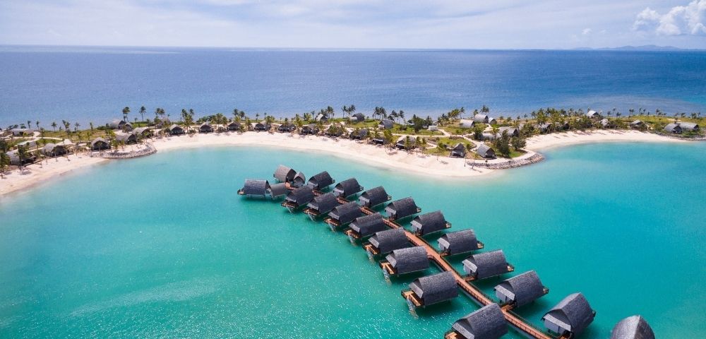 Aerial view of a tropical resort with overwater bungalows and beachfront villas nestled on a white sand island surrounded by turquoise water.