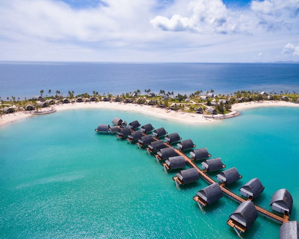 Aerial view of a tropical resort with overwater bungalows and beachfront villas nestled on a white sand island surrounded by turquoise water.