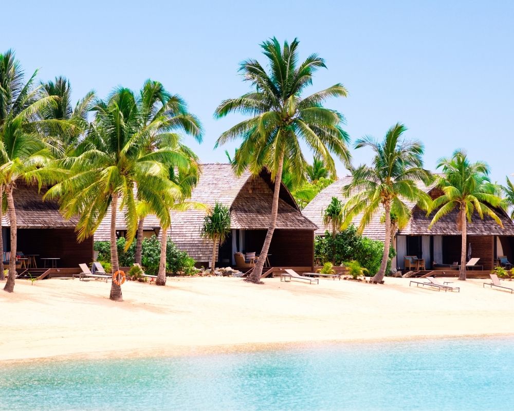 A row of thatched-roof bungalows sits on a sandy beach lined with lush palm trees under a clear blue sky.
