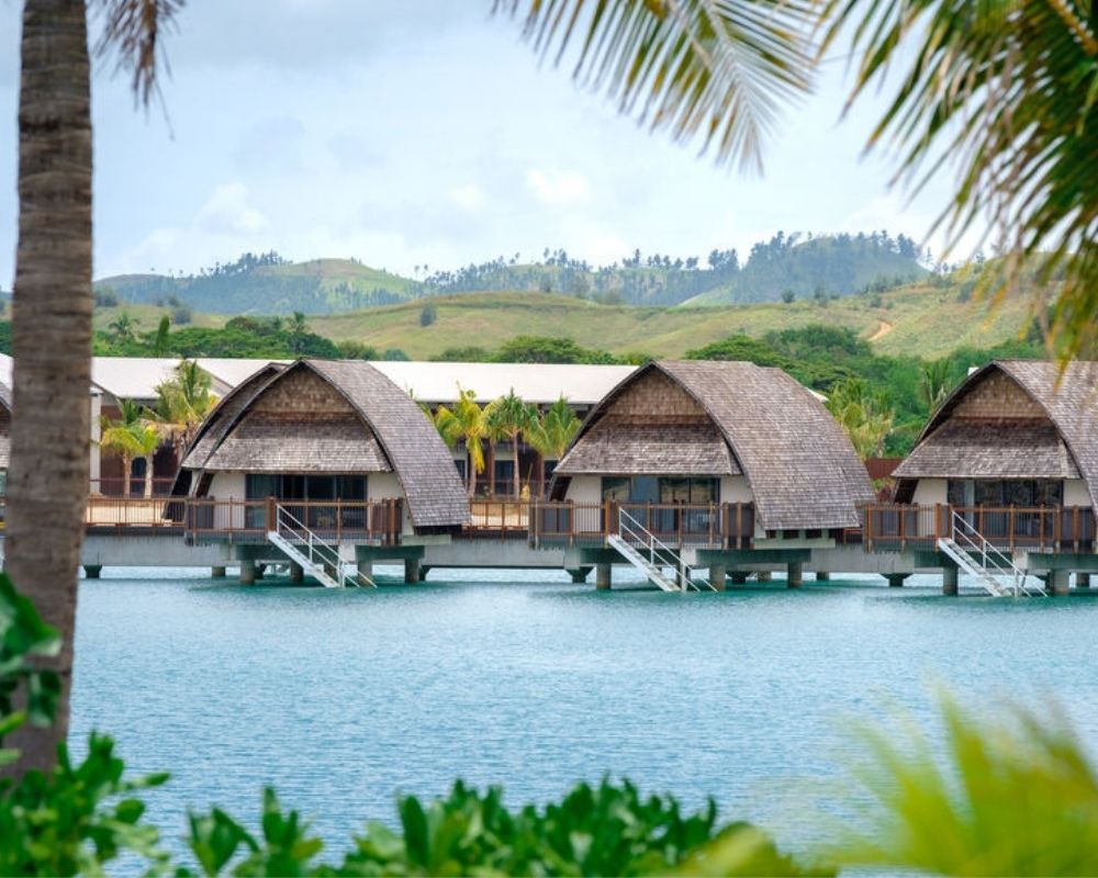 Overwater bungalows with thatched roofs line a tranquil turquoise lagoon, with lush green hills in the background.