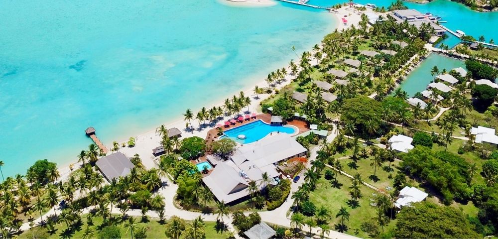 Aerial view of a tropical resort with bungalows, palm trees, a swimming pool, and turquoise water.