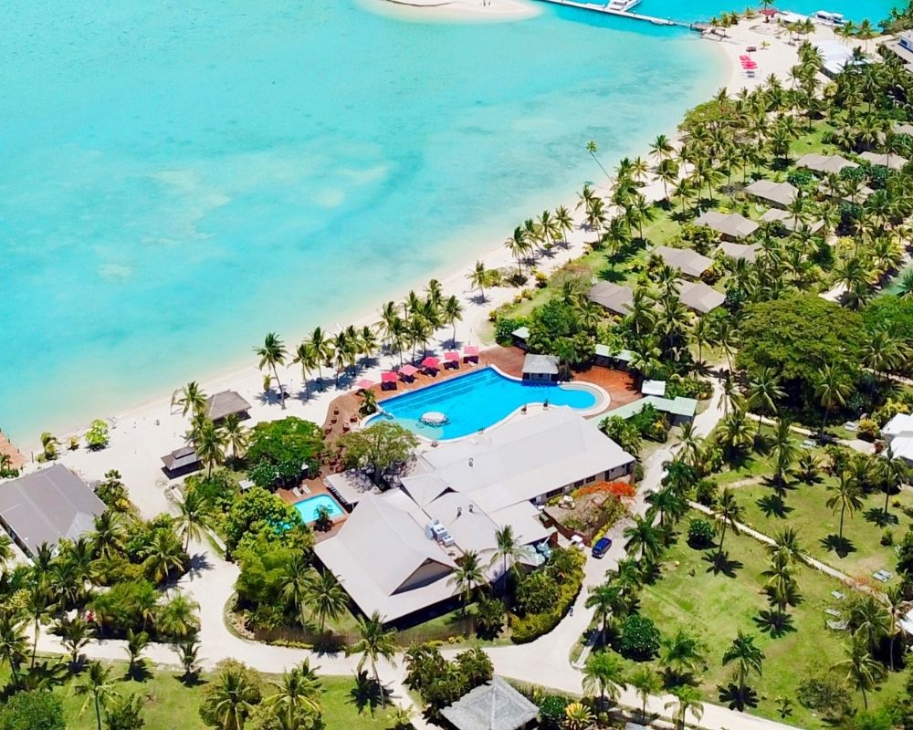 Aerial view of a tropical resort with bungalows, palm trees, a swimming pool, and turquoise water.