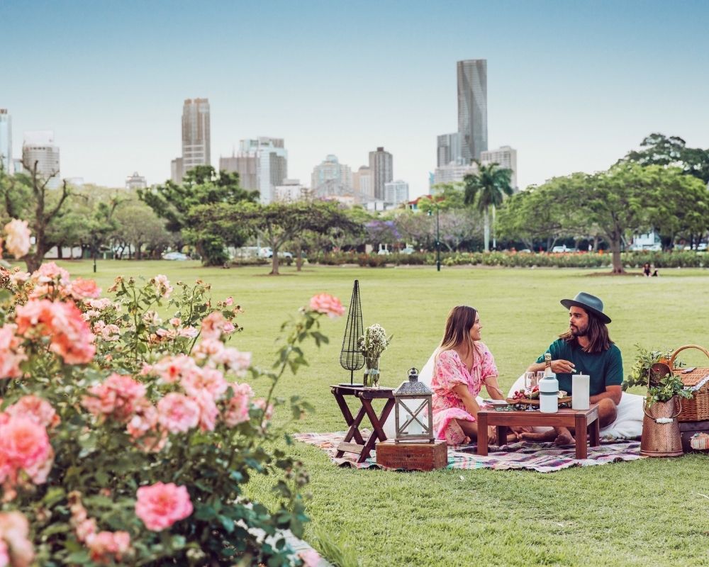 A couple enjoys a picnic on a checkered blanket in a grassy park, with a city skyline visible in the background behind trees and blooming pink roses in the foreground.