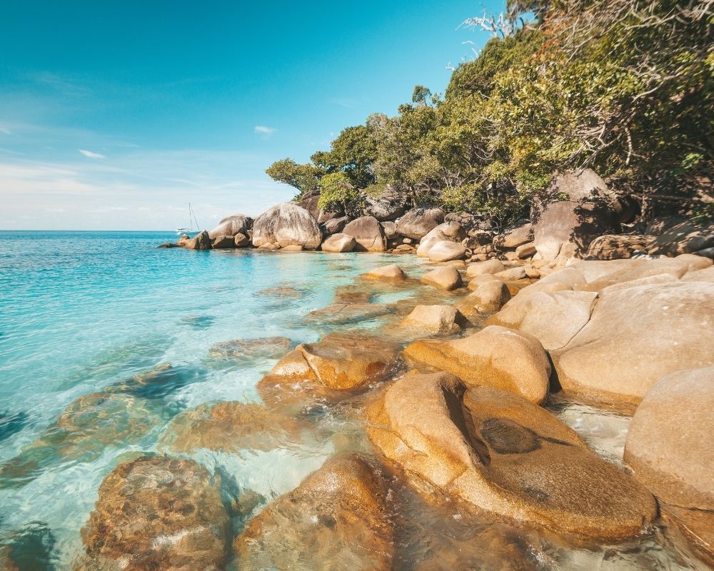 Crystal clear turquoise water laps gently on a rocky shore covered in smooth, tan boulders. Lush green trees and vegetation grow on the slope of a hill in the background, with a sailboat visible in the distance.
