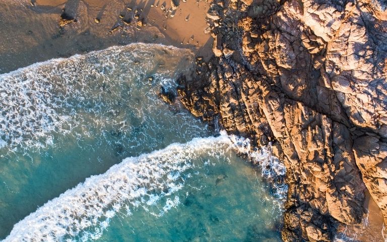 Aerial view of turquoise ocean waves crashing onto a rocky shoreline and sandy beach.
