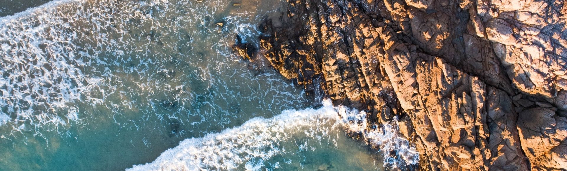 Aerial view of turquoise ocean waves crashing onto a rocky shoreline and sandy beach.