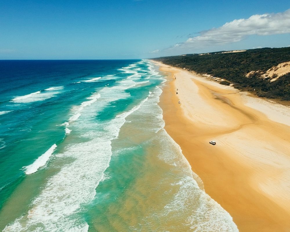 Aerial view of a vast, sandy beach meeting turquoise ocean waves, with lush green vegetation and sand dunes in the background.