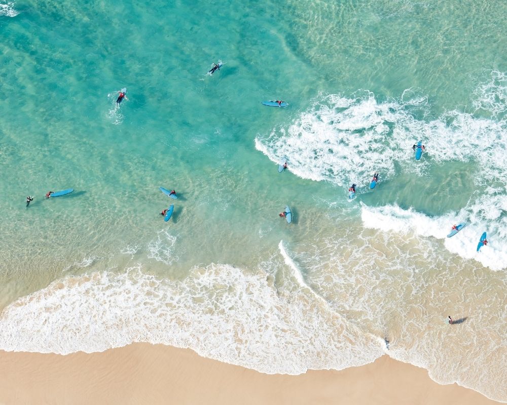 Aerial view of multiple surfers on blue and red surfboards in clear turquoise ocean water with breaking waves and a sandy beach.
